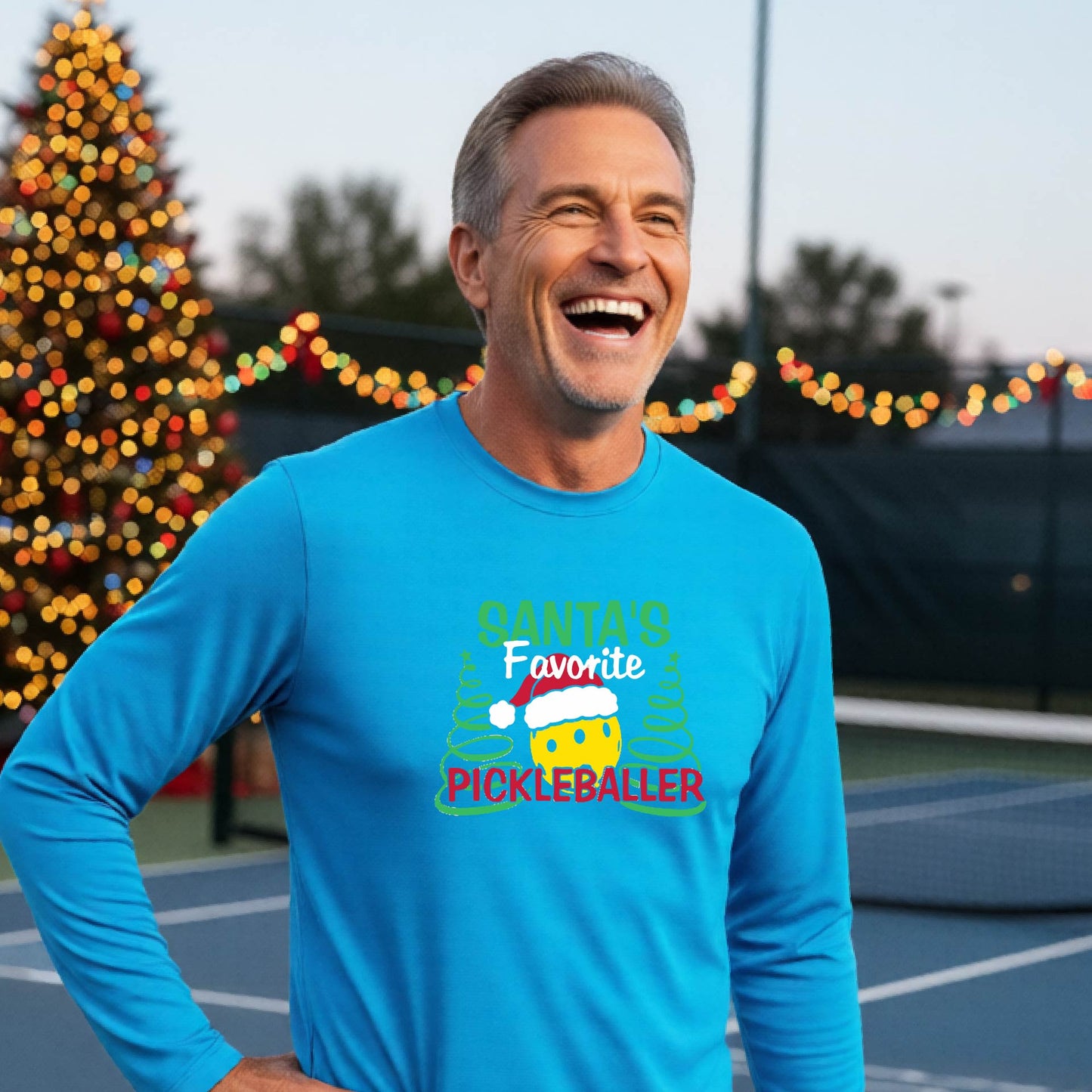 A man wearing a light blue long-sleeve t-shirt with a  pickleball graphic with text "SANTA'S FAVORITE PICKLEBALLER" featuring a bright yellow pickleball set against a blurred background of a pickleball court and a decorated Christmas tree.