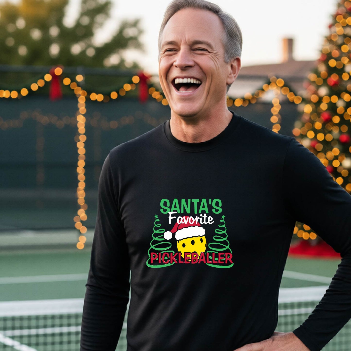 A man wearing a black long-sleeve t-shirt with a  pickleball graphic with text "SANTA'S FAVORITE PICKLEBALLER" featuring a bright yellow pickleball set against a blurred background of a pickleball court and a decorated Christmas tree.