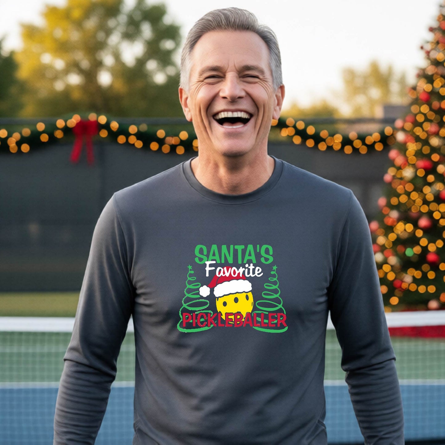 A man wearing a gray long-sleeve t-shirt with a  pickleball graphic with text "SANTA'S FAVORITE PICKLEBALLER" featuring a bright yellow pickleball set against a blurred background of a pickleball court and a decorated Christmas tree.