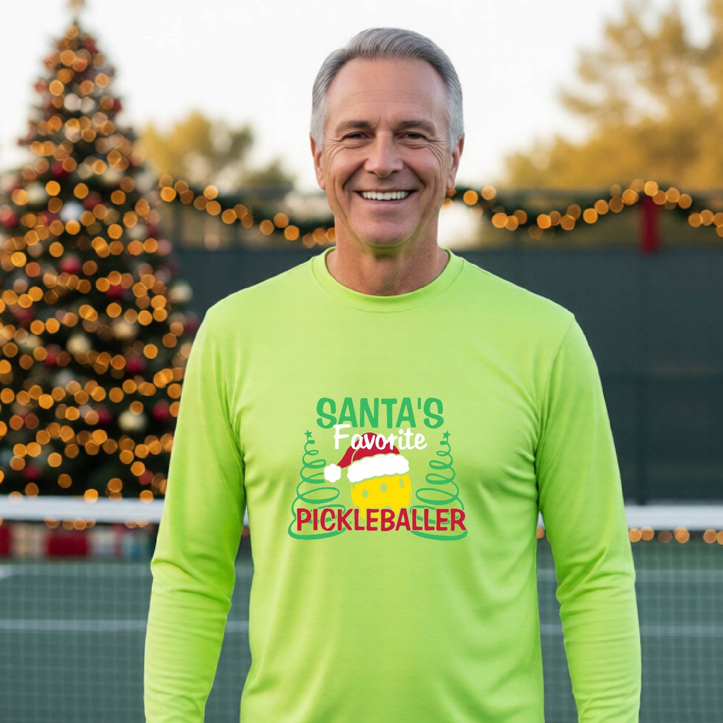 A man wearing a lime green long-sleeve t-shirt with a  pickleball graphic with text "SANTA'S FAVORITE PICKLEBALLER" featuring a bright yellow pickleball set against a blurred background of a pickleball court and a decorated Christmas tree.