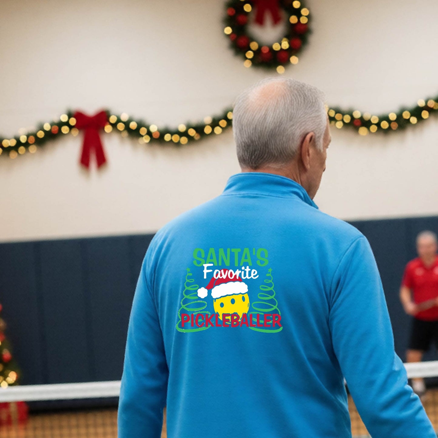 An man viewed from behind wearing a blight blue long-sleeve quarter-zip pullover with a colorful Christmas-themed back graphic that reads 'Santa's Favorite Pickleballer' with a pine tree design. The background shows a pickleball court decorated with holiday garlands, lights, and a wreath.
