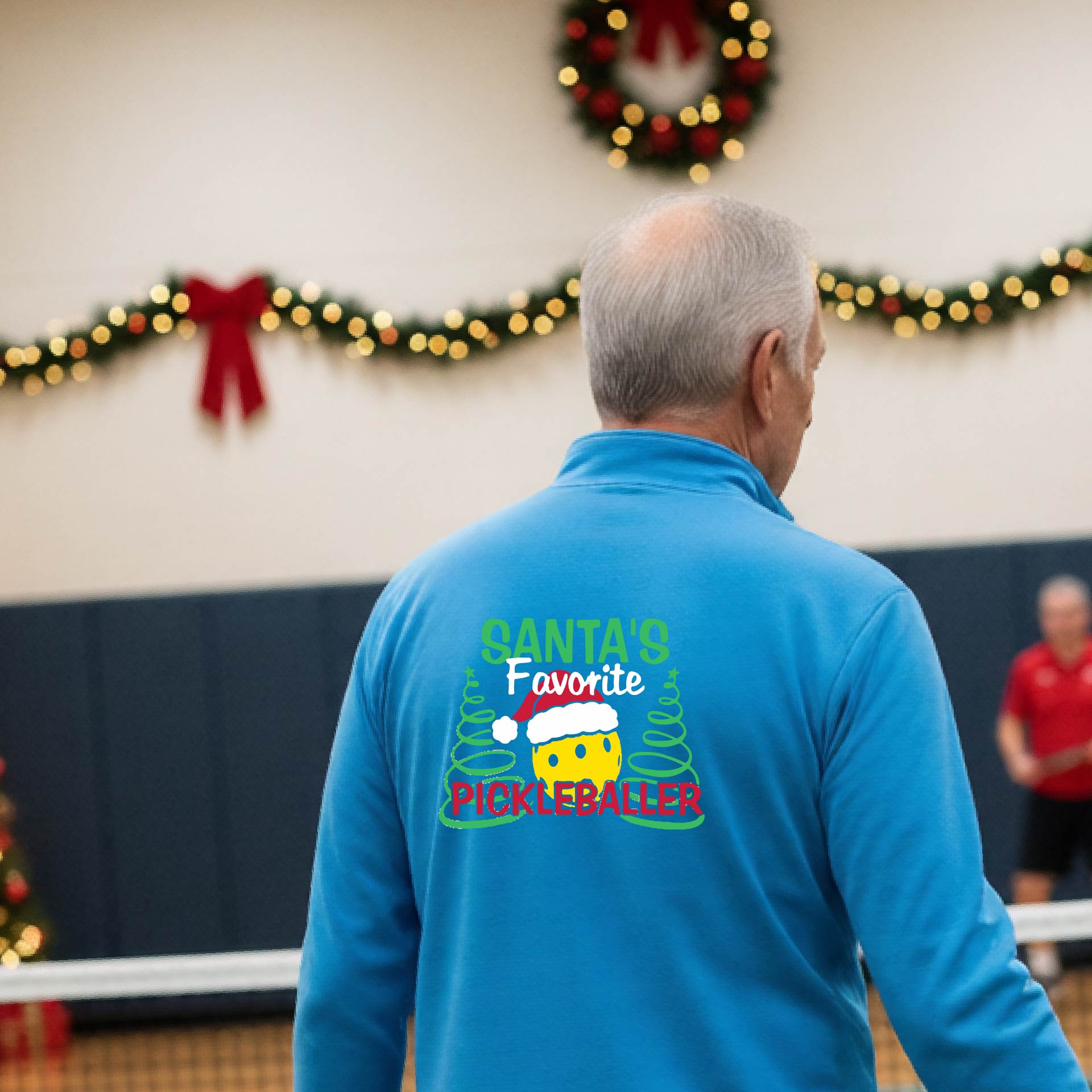 An man viewed from behind wearing a blight blue long-sleeve quarter-zip pullover with a colorful Christmas-themed back graphic that reads 'Santa's Favorite Pickleballer' with a pine tree design. The background shows a pickleball court decorated with holiday garlands, lights, and a wreath.