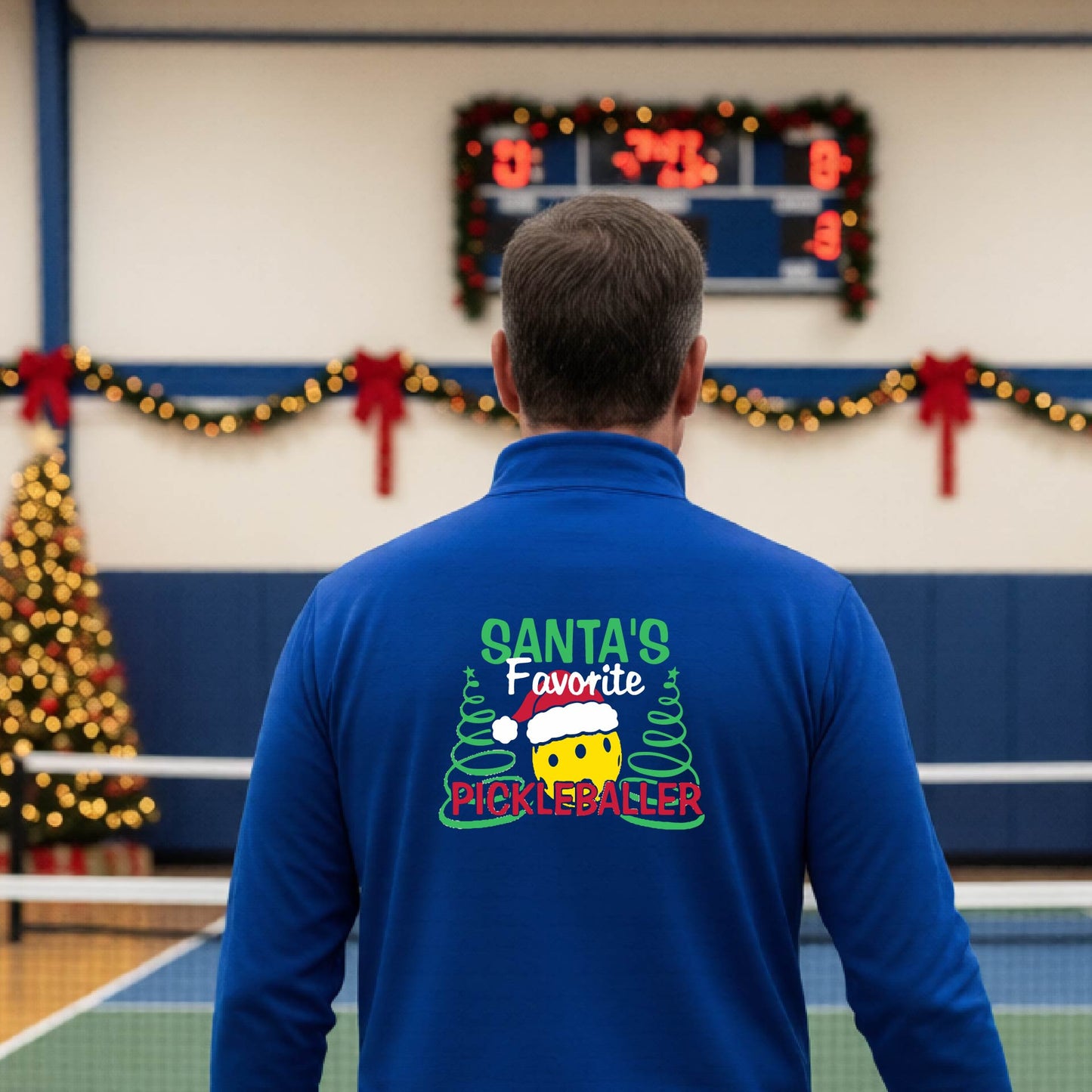 An man viewed from behind wearing a royal blue long-sleeve quarter-zip pullover with a colorful Christmas-themed back graphic that reads 'Santa's Favorite Pickleballer' with a pine tree design. The background shows a pickleball court decorated with holiday garlands, lights, and a wreath.