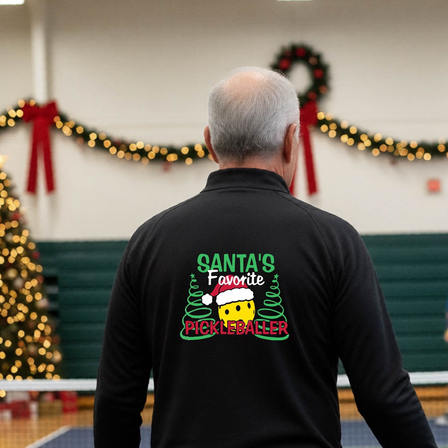 An man viewed from behind wearing a black long-sleeve quarter-zip pullover with a colorful Christmas-themed back graphic that reads 'Santa's Favorite Pickleballer' with a pine tree design. The background shows a pickleball court decorated with holiday garlands, lights, and a wreath.