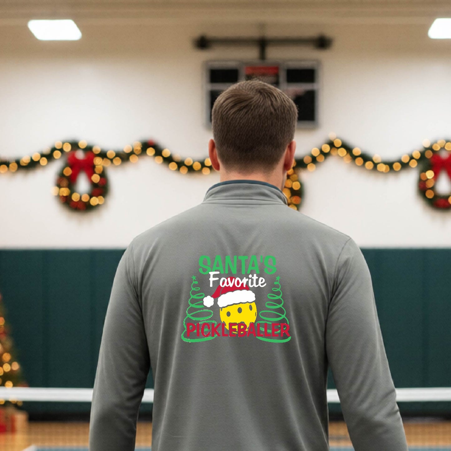 An man viewed from behind wearing a gray long-sleeve quarter-zip pullover with a colorful Christmas-themed back graphic that reads 'Santa's Favorite Pickleballer' with a pine tree design. The background shows a pickleball court decorated with holiday garlands, lights, and a wreath.