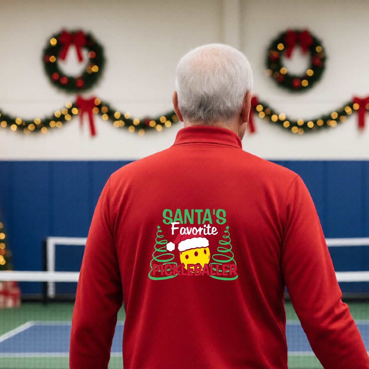 An man viewed from behind wearing a red long-sleeve quarter-zip pullover with a colorful Christmas-themed back graphic that reads 'Santa's Favorite Pickleballer' with a pine tree design. The background shows a pickleball court decorated with holiday garlands, lights, and a wreath.