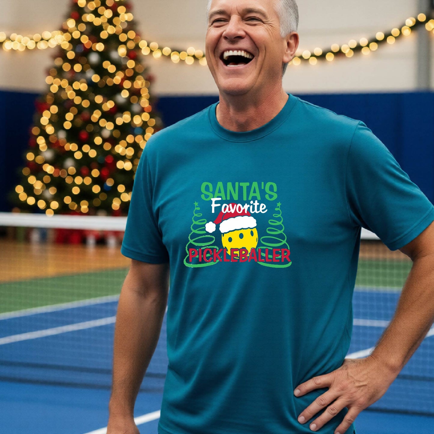 A man wearing a teal short-sleeve t-shirt with a pickleball graphic with text "SANTA'S FAVORITE PICKLEBALLER" featuring a bright yellow pickleball set against a blurred background of a pickleball court and a decorated Christmas tree.