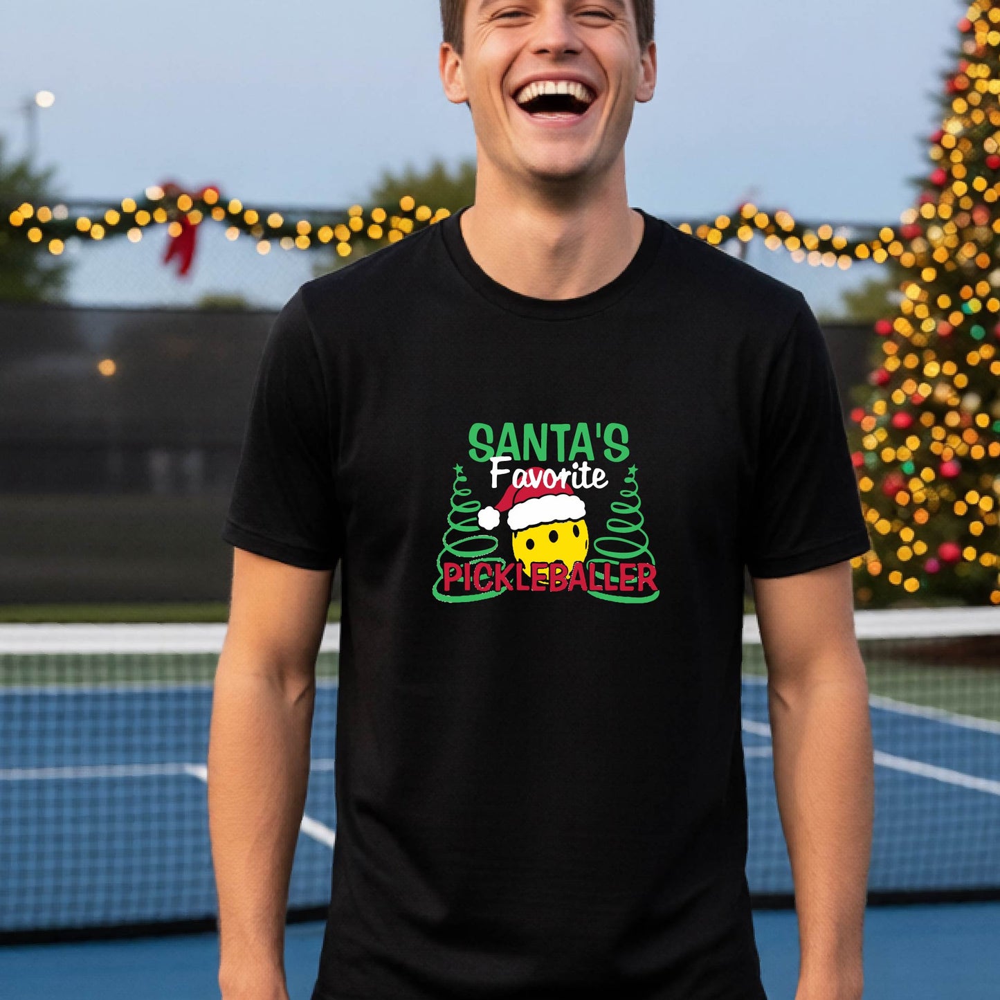 A man wearing a black short-sleeve t-shirt with a pickleball graphic with text "SANTA'S FAVORITE PICKLEBALLER" featuring a bright yellow pickleball set against a blurred background of a pickleball court and a decorated Christmas tree.