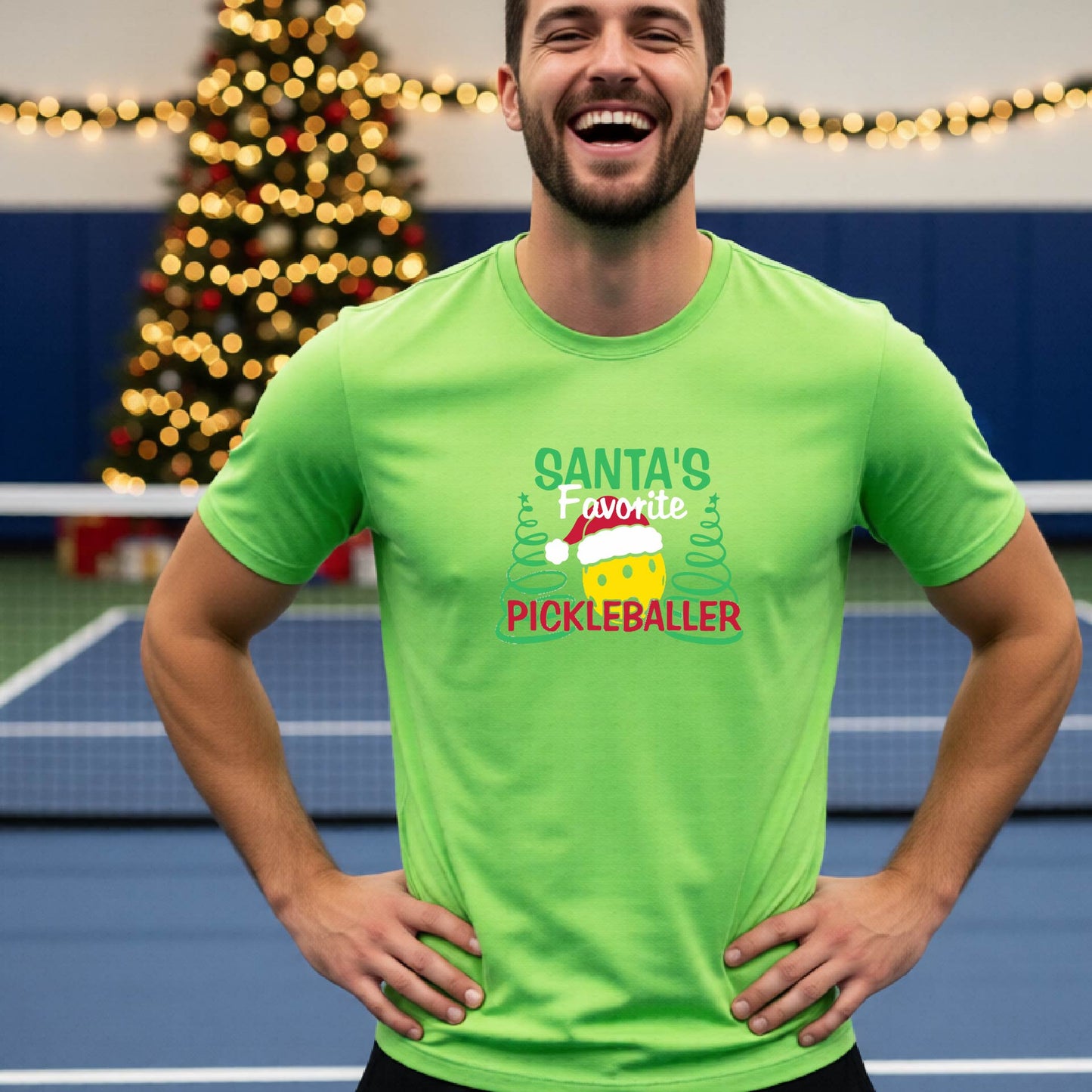 A man wearing a lime green short-sleeve t-shirt with a pickleball graphic with text "SANTA'S FAVORITE PICKLEBALLER" featuring a bright yellow pickleball set against a blurred background of a pickleball court and a decorated Christmas tree.