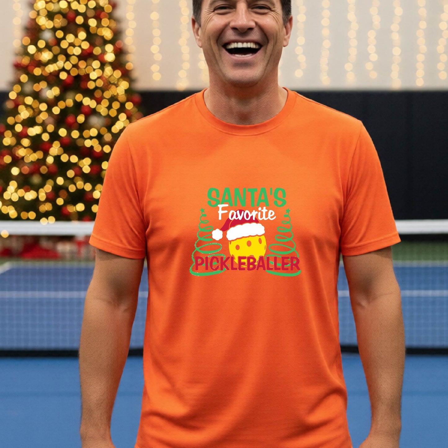 A man wearing an orange short-sleeve t-shirt with a pickleball graphic with text "SANTA'S FAVORITE PICKLEBALLER" featuring a bright yellow pickleball set against a blurred background of a pickleball court and a decorated Christmas tree.