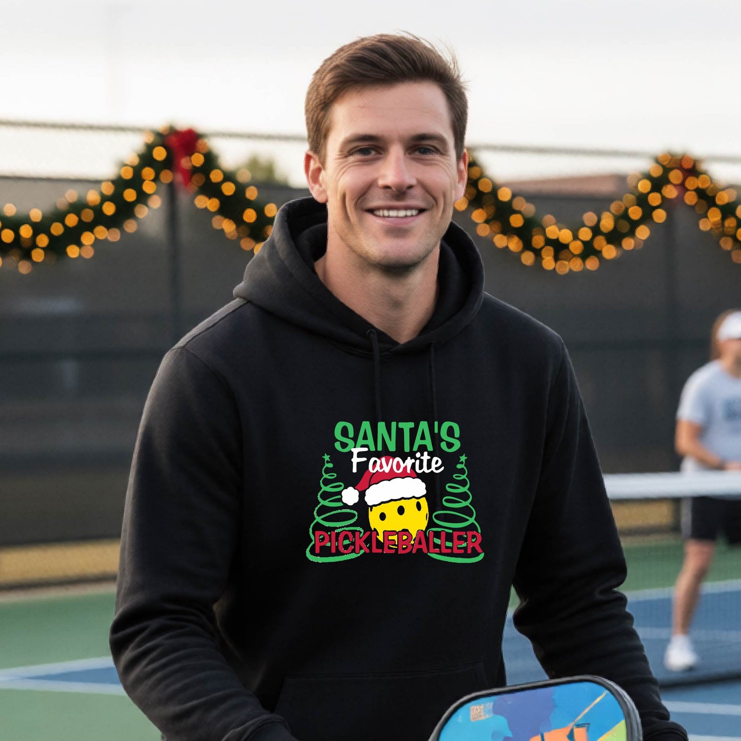 A smiling man wearing a black hoodie with a festive graphic that reads 'Santa's Favorite Pickleballer' stands on an pickleball court. The graphic features a yellow pickleball character wearing a Santa hat, green pine trees, and holiday garlands in the background.