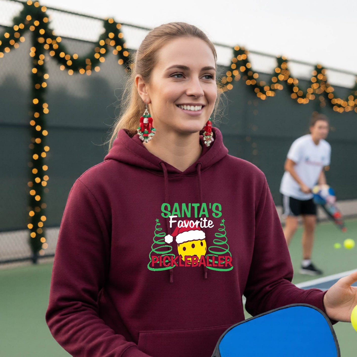 A smiling woman wearing a maroon hoodie with a festive graphic that reads 'Santa's Favorite Pickleballer' stands on an pickleball court. The graphic features a yellow pickleball character wearing a Santa hat, green pine trees, and holiday garlands in the background.