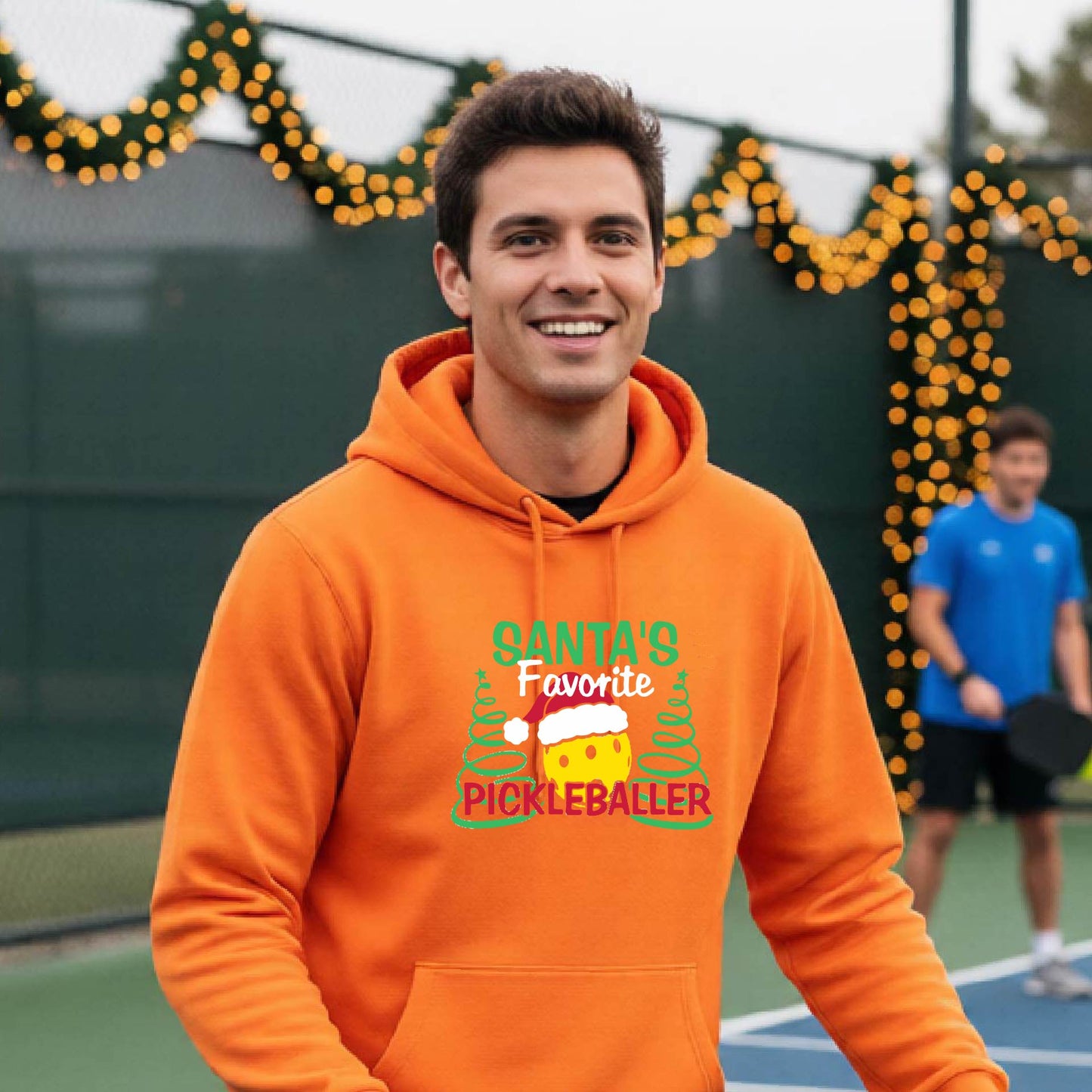 A smiling man wearing a bright orange hoodie with a festive graphic that reads 'Santa's Favorite Pickleballer' stands on an pickleball court. The graphic features a yellow pickleball character wearing a Santa hat, green pine trees, and holiday garlands in the background.