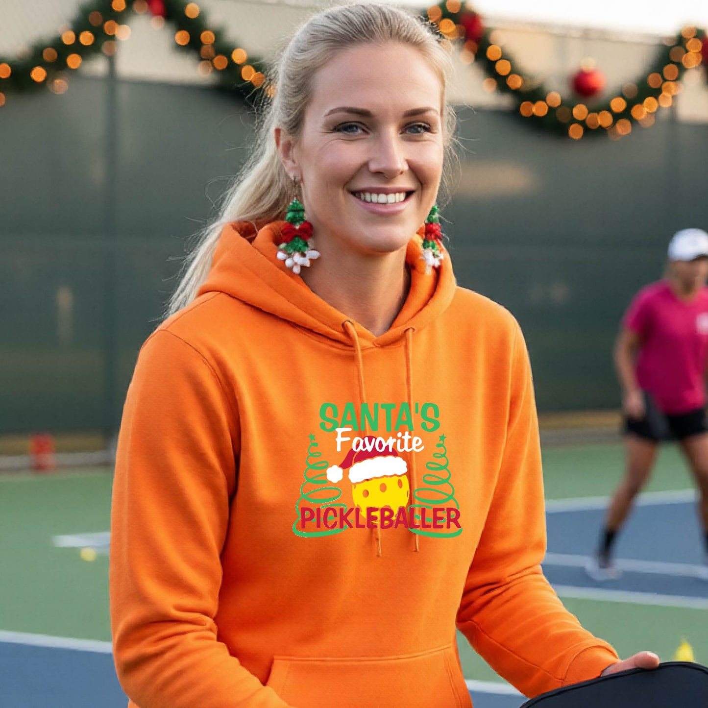 A smiling woman wearing a bright orange hoodie with a festive graphic that reads 'Santa's Favorite Pickleballer' stands on an pickleball court. The graphic features a yellow pickleball character wearing a Santa hat, green pine trees, and holiday garlands in the background.