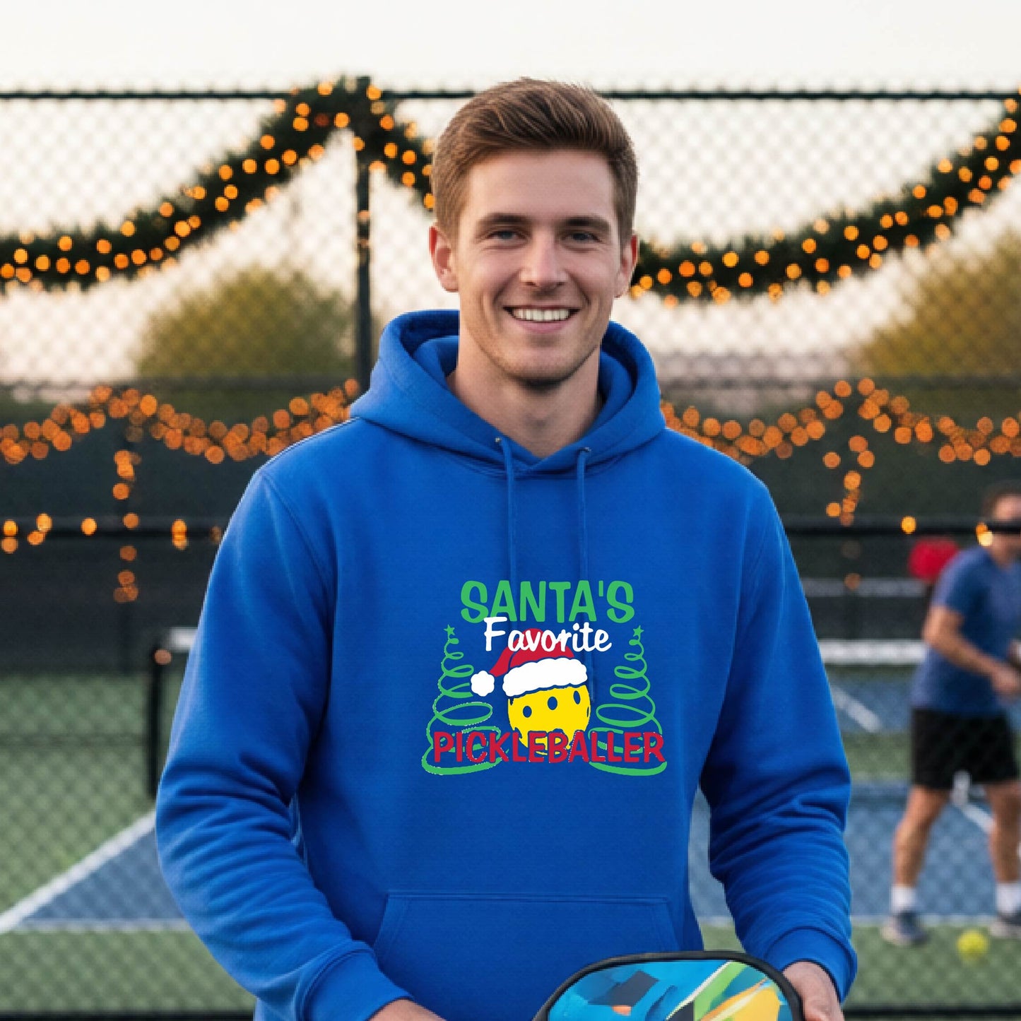 A smiling man wearing a bright blue hoodie with a festive graphic that reads 'Santa's Favorite Pickleballer' stands on an pickleball court. The graphic features a yellow pickleball character wearing a Santa hat, green pine trees, and holiday garlands in the background.