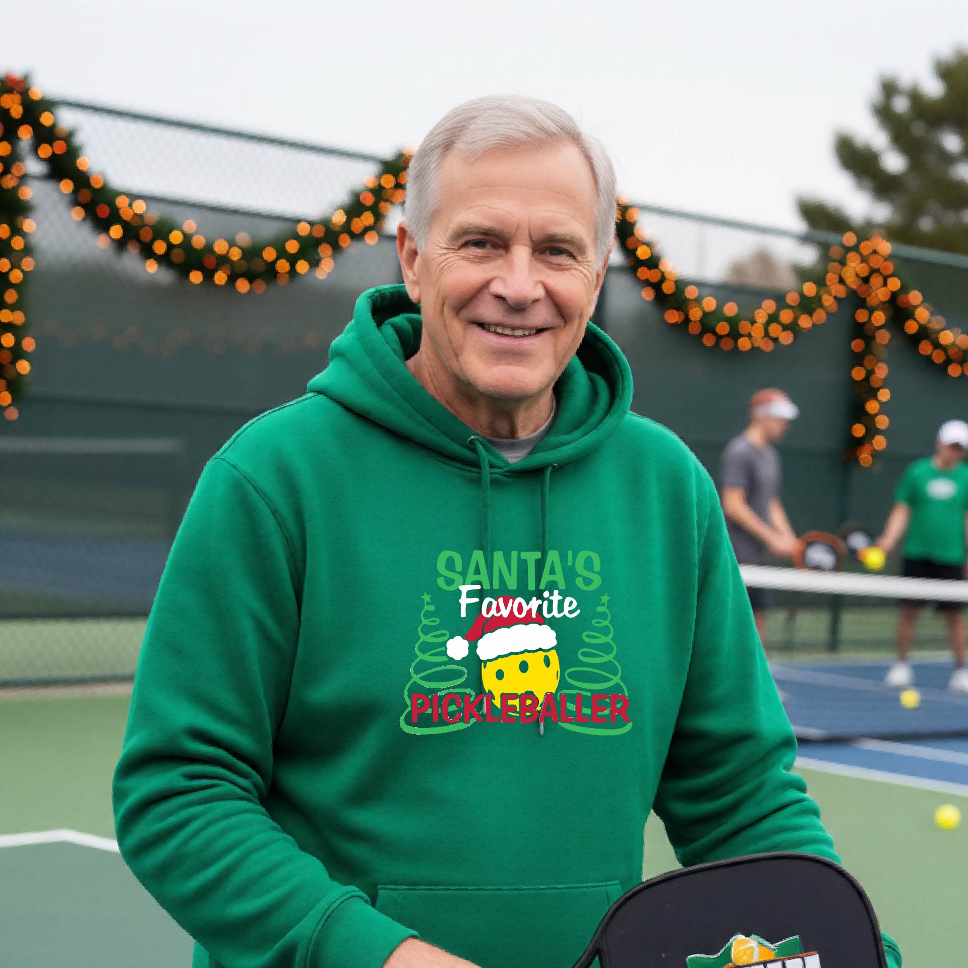 A smiling man wearing a green hoodie with a festive graphic that reads 'Santa's Favorite Pickleballer' stands on an pickleball court. The graphic features a yellow pickleball character wearing a Santa hat, green pine trees, and holiday garlands in the background.
