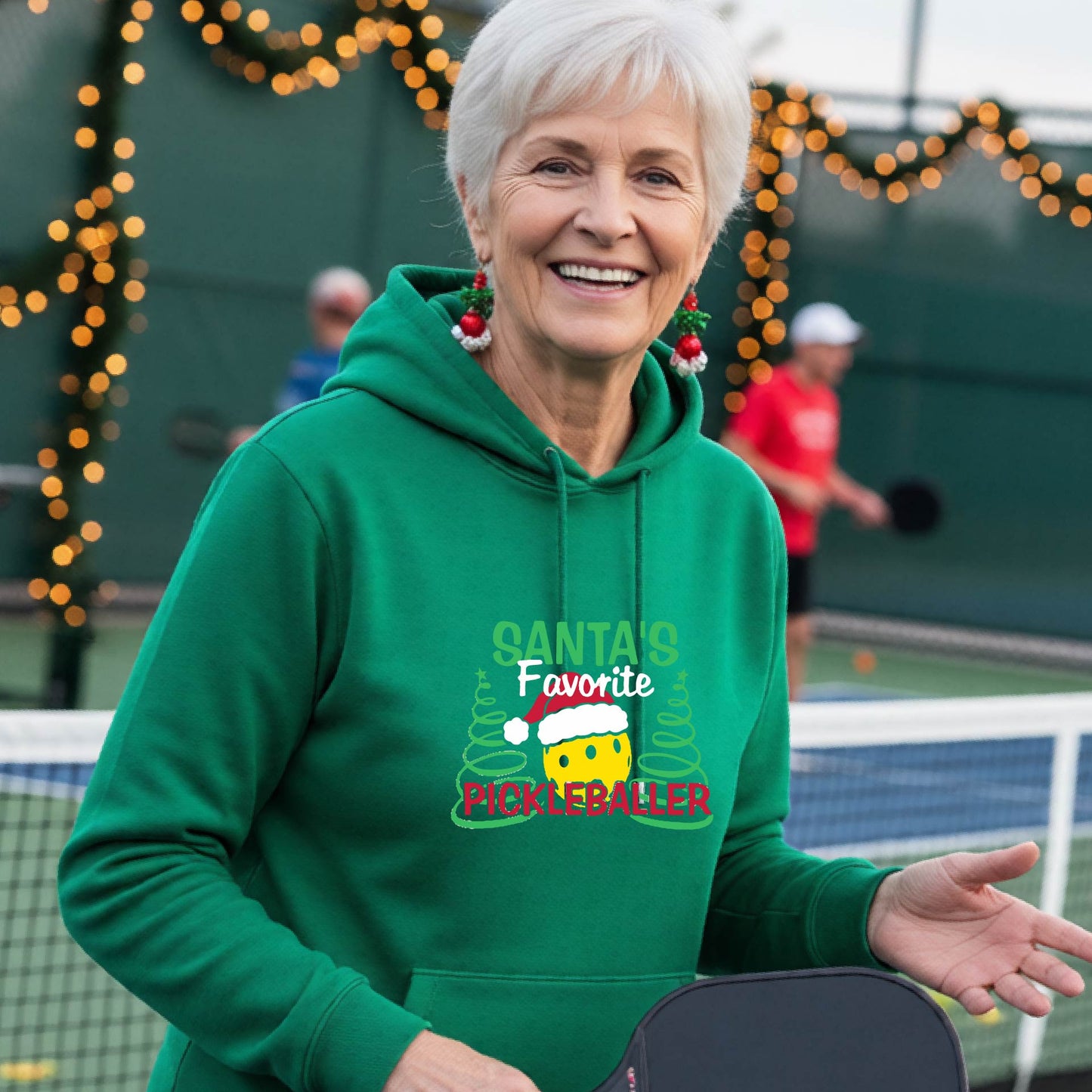 A smiling woman wearing a bright green hoodie with a festive graphic that reads 'Santa's Favorite Pickleballer' stands on an pickleball court. The graphic features a yellow pickleball character wearing a Santa hat, green pine trees, and holiday garlands in the background.