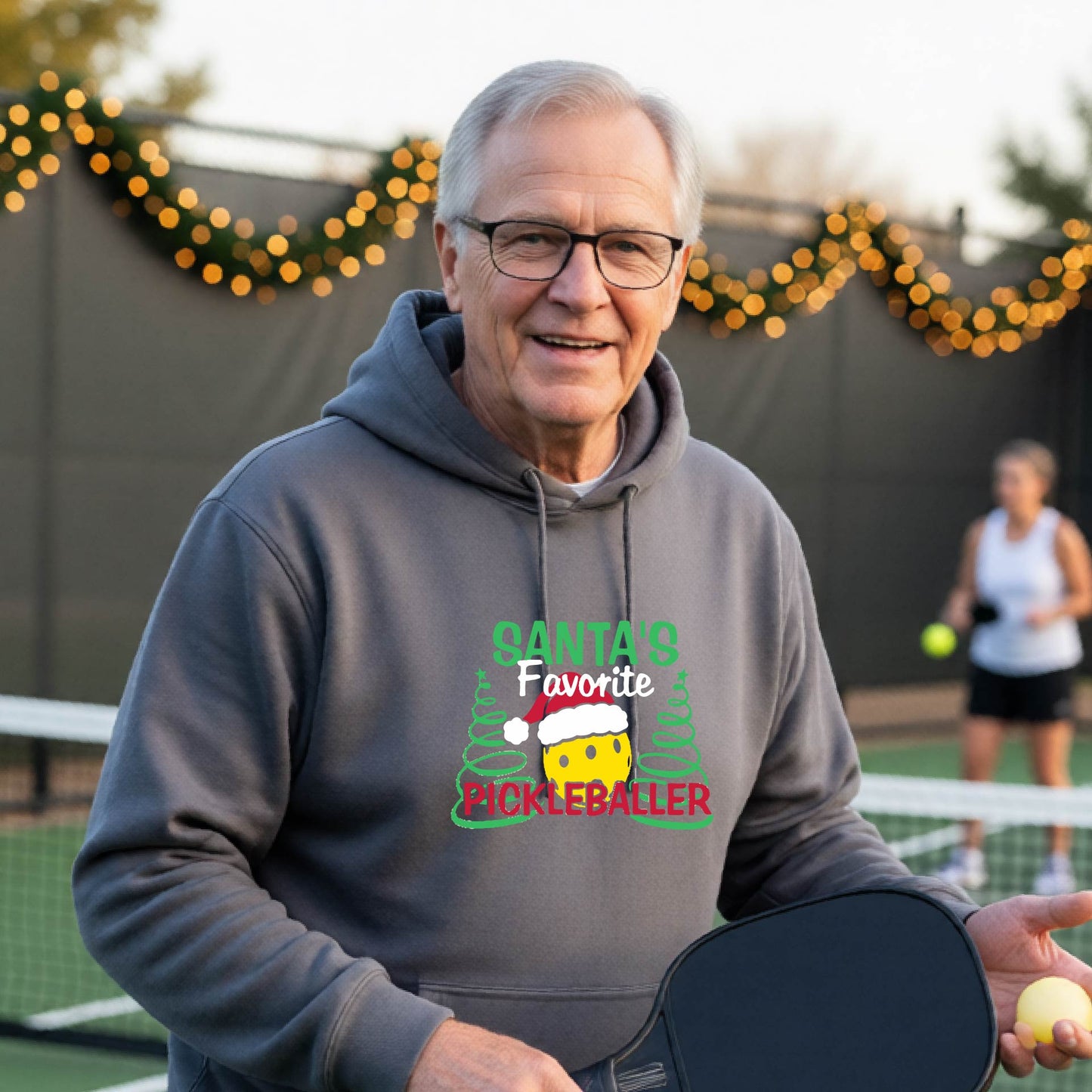 A smiling man wearing a gray hoodie with a festive graphic that reads 'Santa's Favorite Pickleballer' stands on an pickleball court. The graphic features a yellow pickleball character wearing a Santa hat, green pine trees, and holiday garlands in the background.