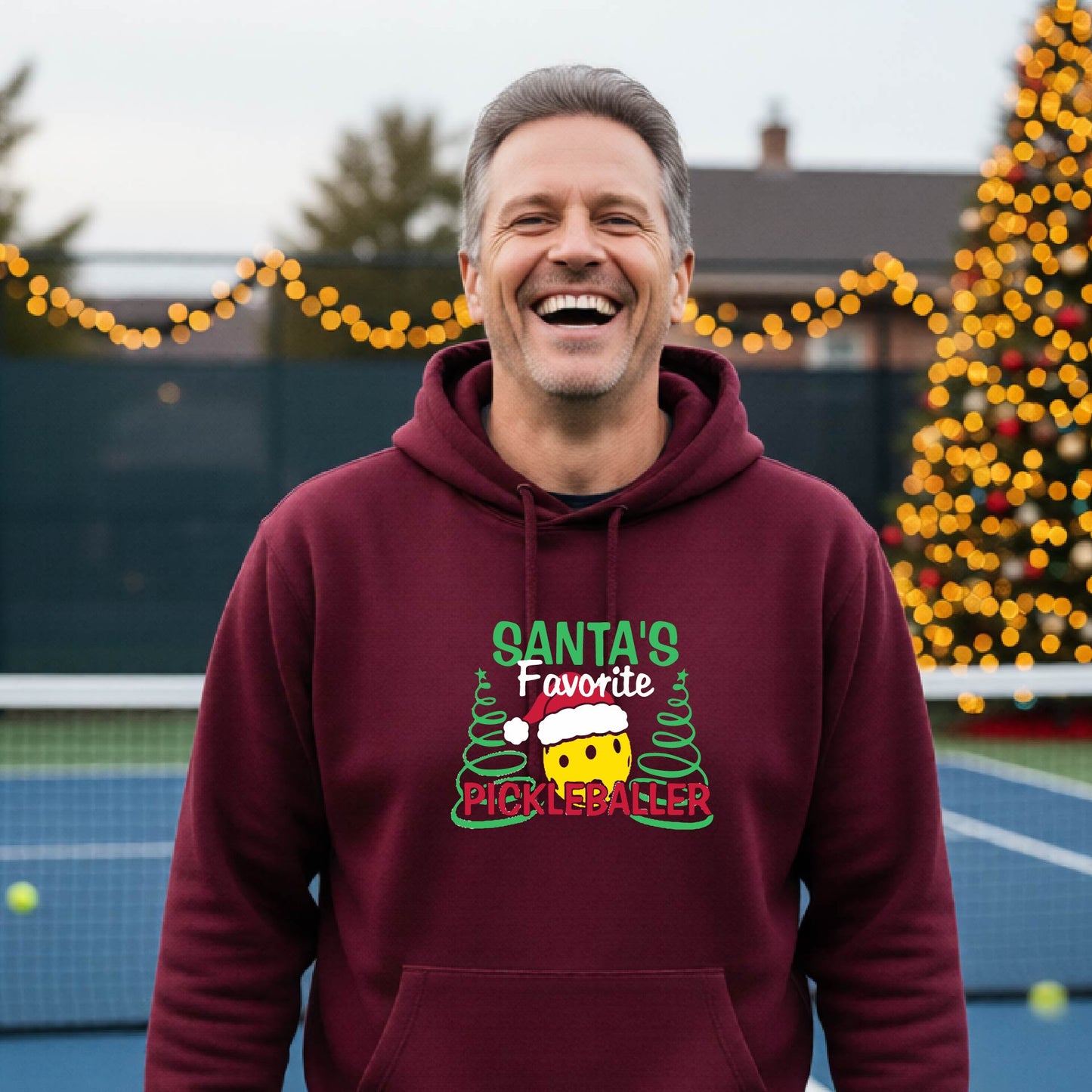 A smiling man wearing a maroon hoodie with a festive graphic that reads 'Santa's Favorite Pickleballer' stands on an pickleball court. The graphic features a yellow pickleball character wearing a Santa hat, green pine trees, and holiday garlands in the background.