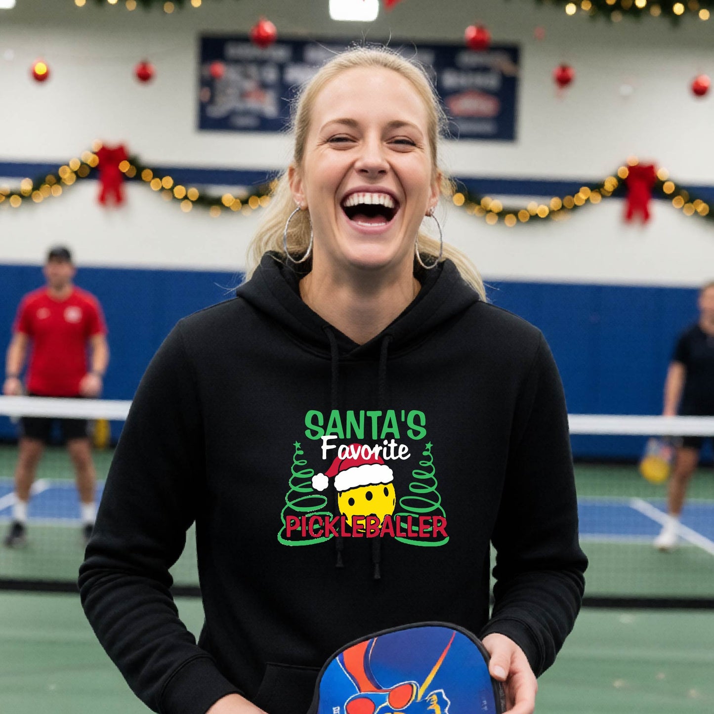 A black hooded sweatshirt featuring a pickleball graphic with the text "SANTA'S FAVORITE PICKLEBALL." and a Christmas 