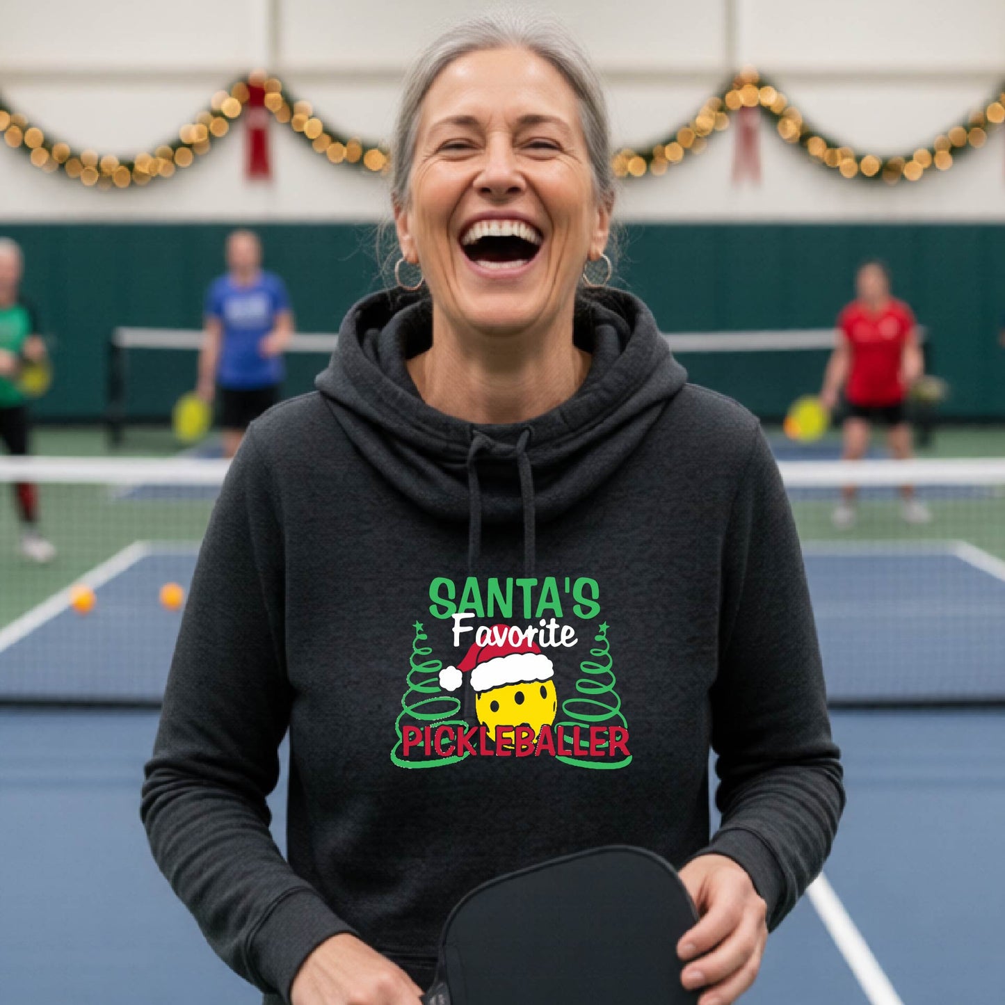 A black cowl-neck hooded sweatshirt featuring a pickleball graphic and text "SANTA'S FAVORITE PICKLEBALLER" and Christmas themed background. 