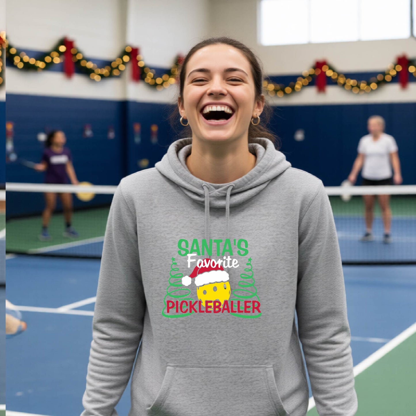 A grey cowl-neck hooded sweatshirt featuring a pickleball graphic and text "SANTA'S FAVORITE PICKLEBALLER" and Christmas themed background. 