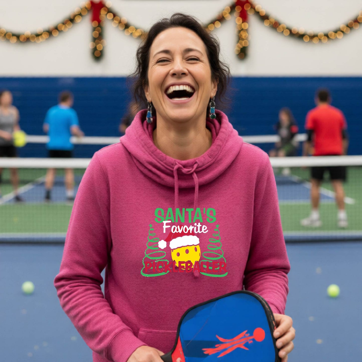 A pink cowl-neck hooded sweatshirt featuring a pickleball graphic and text "SANTA'S FAVORITE PICKLEBALLER" and Christmas themed background. 