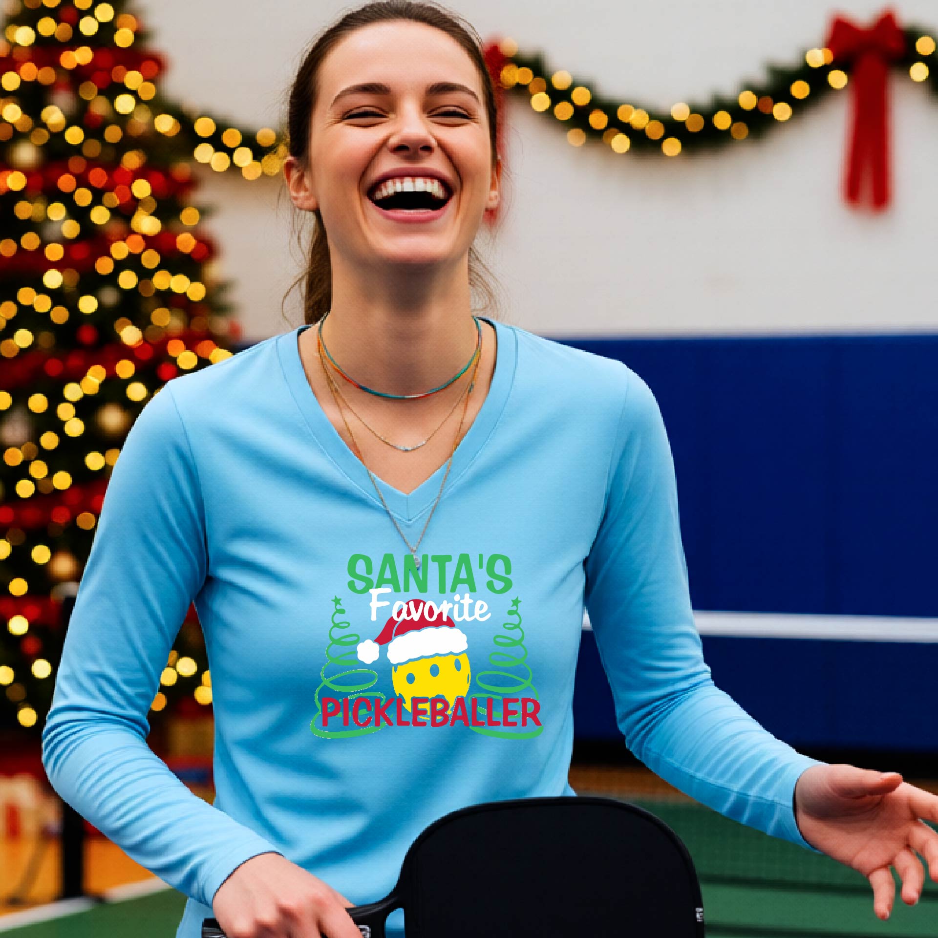 A woman wearing a light blue long-sleeve V-neck t-shirt with a colorful graphic that reads 'Santa's Favorite Pickleballer' with a yellow pickleball character in a Santa hat, set against a blurred background of a Christmas tree and lights.
