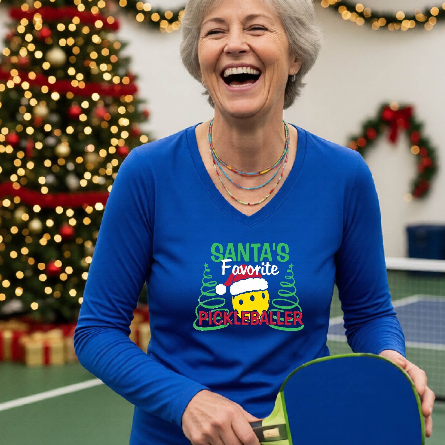 A woman wearing a royal blue long-sleeve V-neck t-shirt with a colorful graphic that reads 'Santa's Favorite Pickleballer' with a yellow pickleball character in a Santa hat, set against a blurred background of a Christmas tree and lights.