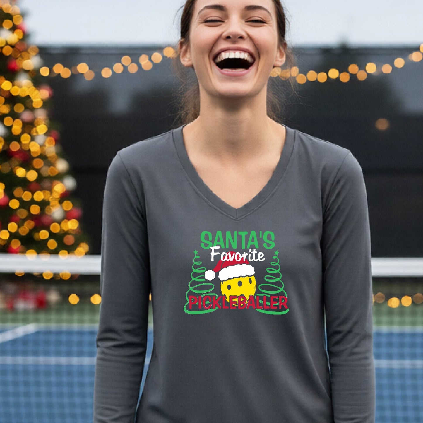 A woman wearing a dark gray long-sleeve V-neck t-shirt with a colorful graphic that reads 'Santa's Favorite Pickleballer' with a yellow pickleball character in a Santa hat, set against a blurred background of a Christmas tree and lights.