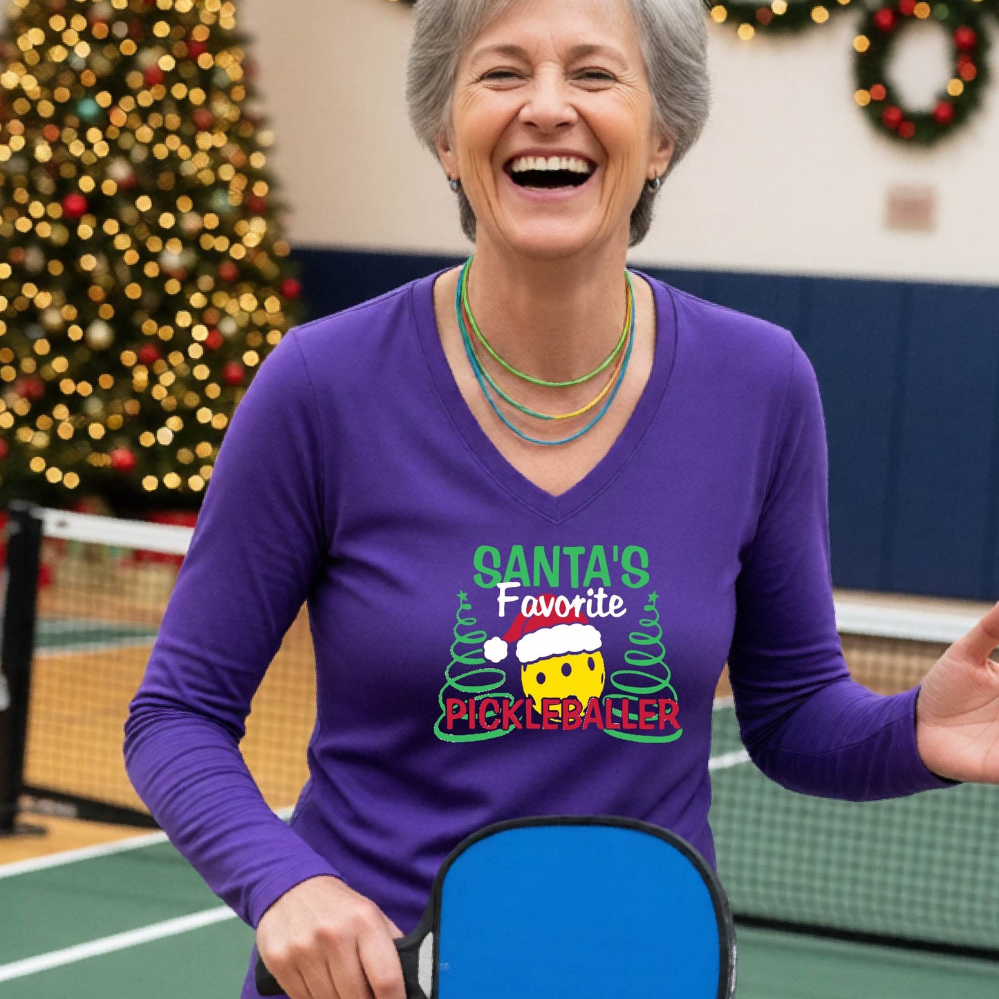 A woman wearing a purple long-sleeve V-neck t-shirt with a colorful graphic that reads 'Santa's Favorite Pickleballer' with a yellow pickleball character in a Santa hat, set against a blurred background of a Christmas tree and lights.