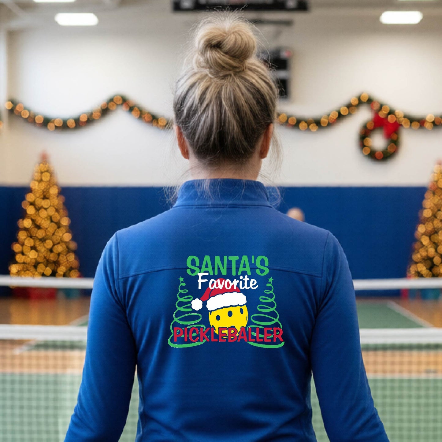 A woman viewed from behind wearing a royal blue long-sleeve athletic shirt with a festive Christmas-themed graphic on the back. The graphic reads 'SANTA'S Favorite PICKLEBALLER' in green, white, and red text, featuring a yellow smiley face wearing a Santa hat between two green pine trees.