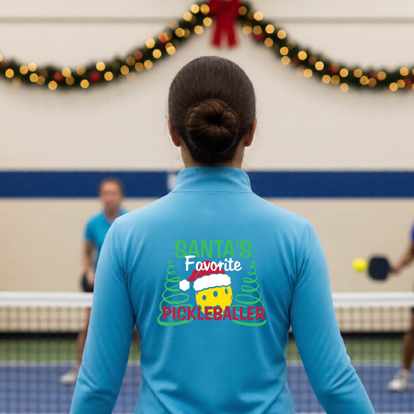 A woman viewed from behind wearing a bright blue long-sleeve athletic top with a large Christmas-themed graphic on the back that reads 'Santa's Favorite Pickleballer' with a smiling emoji wearing a Santa hat and pine trees. The photo is taken in an indoor pickleball court with holiday garlands visible in the background.