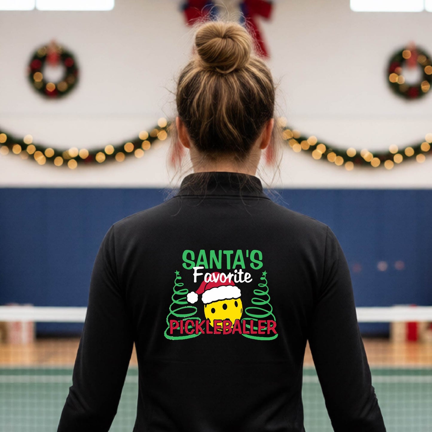 A person viewed from behind wearing a black long-sleeve shirt with a colorful Christmas-themed graphic on the back that reads 'Santa's Favorite Pickleballer' with a yellow smiley face, green trees, and a red Santa hat.