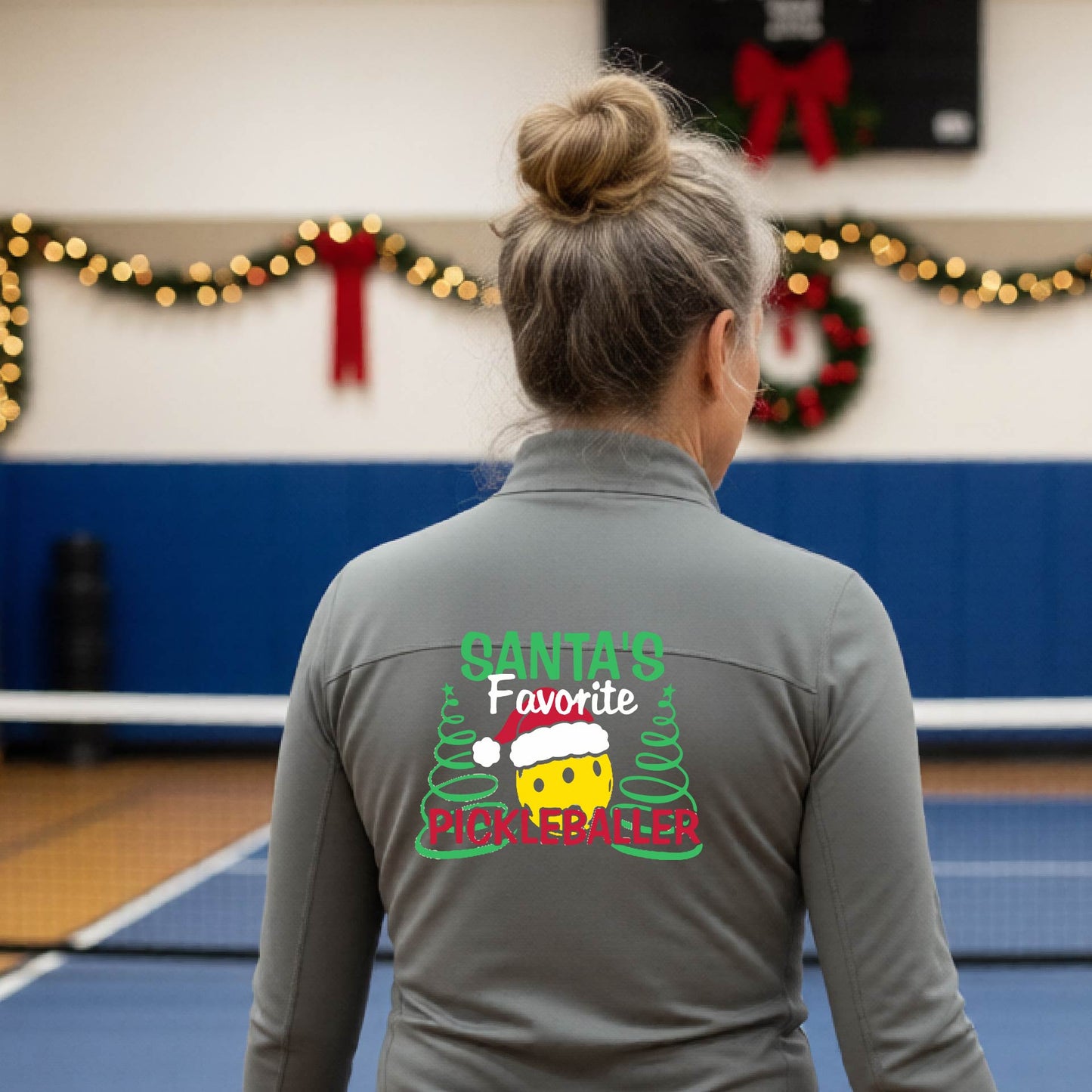 A woman viewed from behind wearing a gray long-sleeve athletic shirt with a colorful Christmas-themed graphic on the back that reads 'Santa's Favorite Pickleballer' with a yellow face and green/red details. She is standing on an indoor pickleball court with holiday decorations visible in the background.