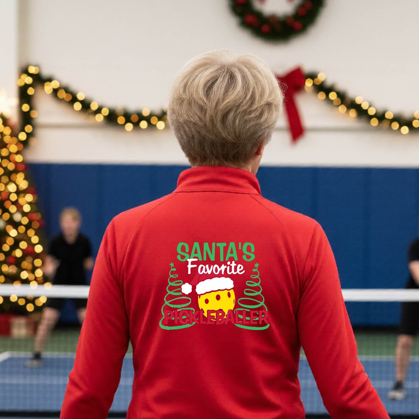 A person viewed from behind wearing a bright red long-sleeve athletic jacket in a holiday-themed indoor sports hall. The back of the jacket features a graphic with the text 'SANTA'S Favorite PICKLEBALLER' and a yellow pickleball character between green Christmas trees.