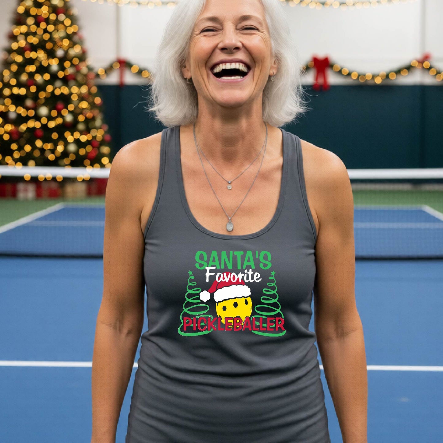 A smiling woman stands on a blue indoor pickleball court wearing a dark gray racerback tank top with a colorful graphic that reads 'Santa's Favorite Pickleballer' with a cartoon pickleball and pine trees.