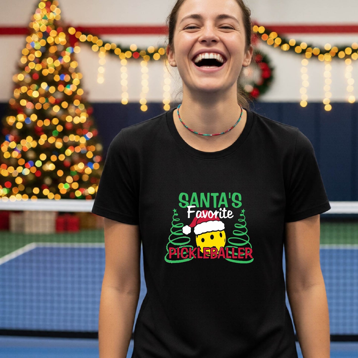 A woman laughing while wearing a black graphic t-shirt that reads 'Santa's Favorite Pickleballer' with a festive pickleball graphic. The background shows a holiday-themed indoor setting with a Christmas tree, and stockings.