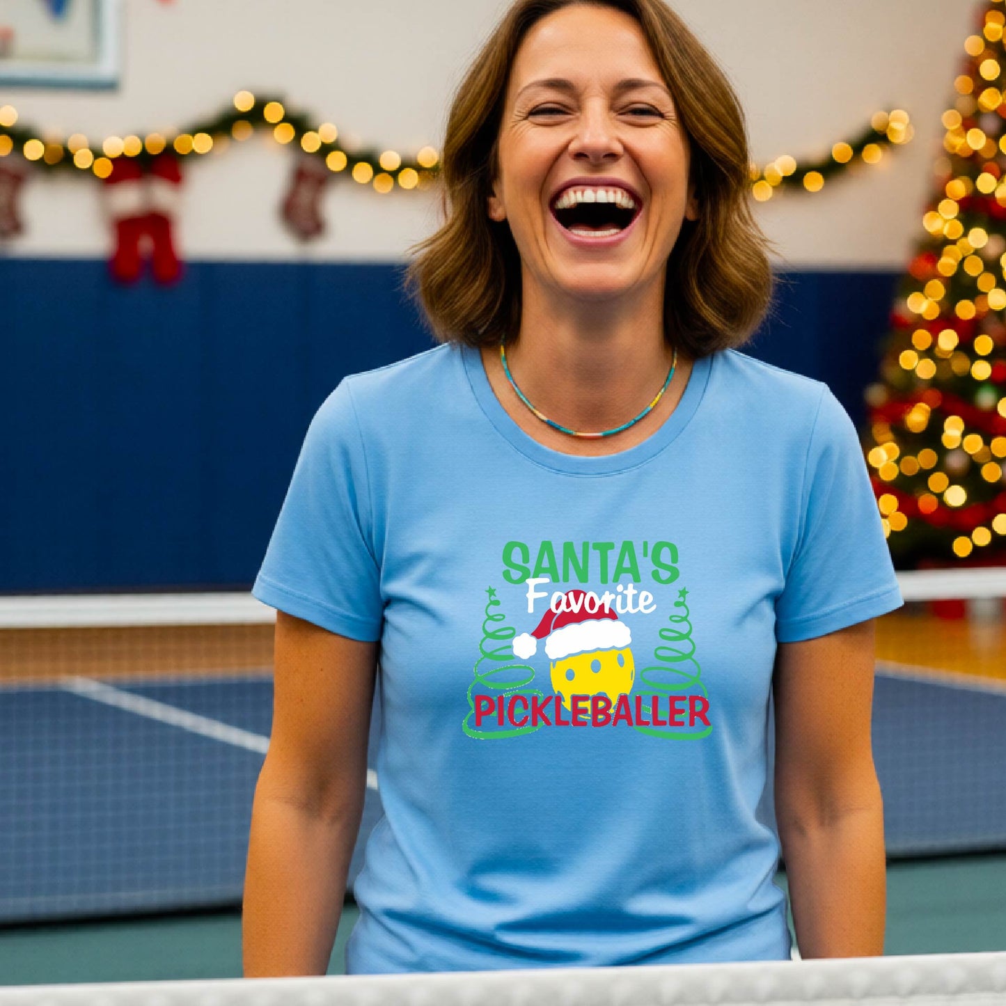 A woman laughing while wearing a light blue graphic t-shirt that reads 'Santa's Favorite Pickleballer' with a festive pickleball graphic. The background shows a holiday-themed indoor setting with a Christmas tree, and stockings.
