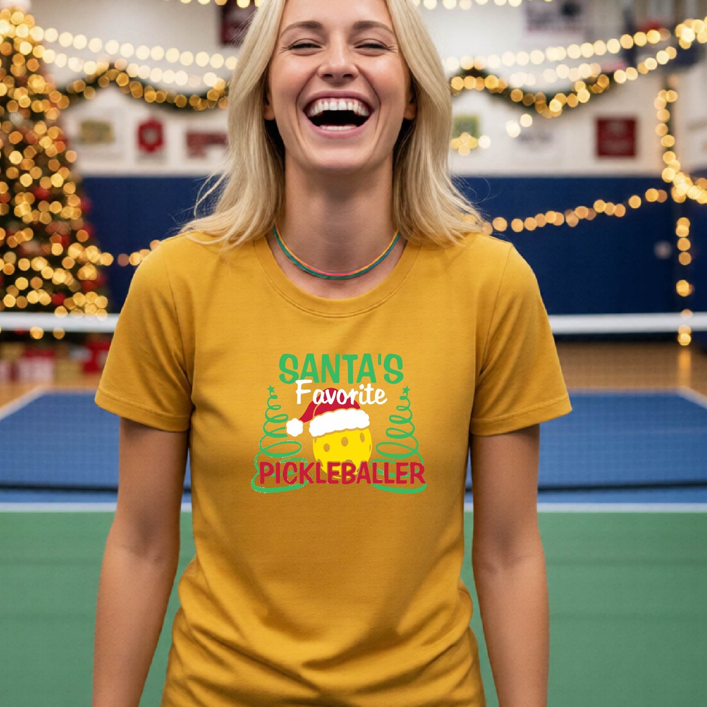 A woman laughing while wearing a mustard yellow gold graphic t-shirt that reads 'Santa's Favorite Pickleballer' with a festive pickleball graphic. The background shows a holiday-themed indoor setting with a Christmas tree, and stockings.