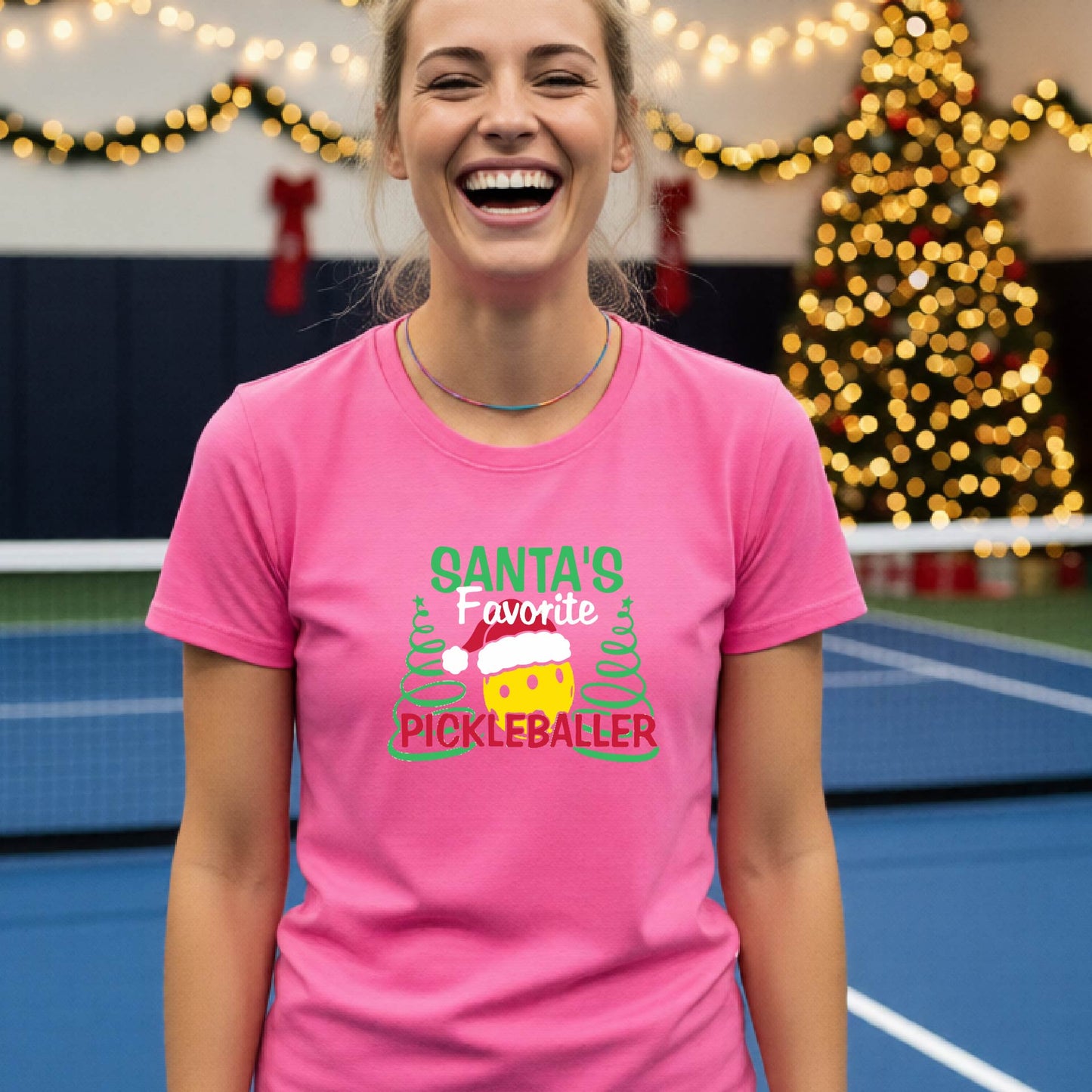 A woman laughing while wearing a bright pink graphic t-shirt that reads 'Santa's Favorite Pickleballer' with a festive pickleball graphic. The background shows a holiday-themed indoor setting with a Christmas tree, and stockings.