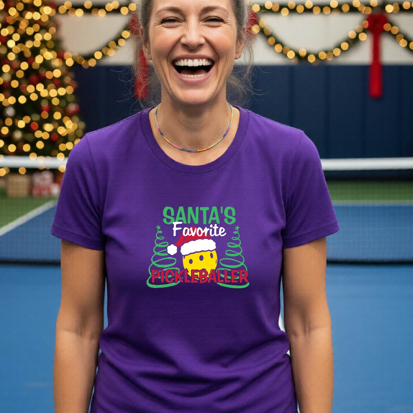 A woman laughing while wearing a purple graphic t-shirt that reads 'Santa's Favorite Pickleballer' with a festive pickleball graphic. The background shows a holiday-themed indoor setting with a Christmas tree, and stockings.