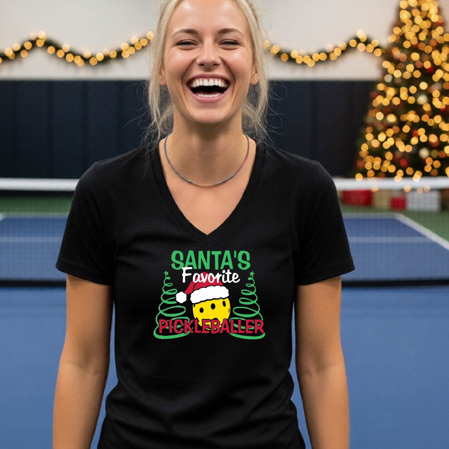 A woman smiling in a gym wearing a black V-neck t-shirt with a 'Santa's Favorite Pickleballer' graphic on the front, with holiday decorations and a Christmas tree in the background.