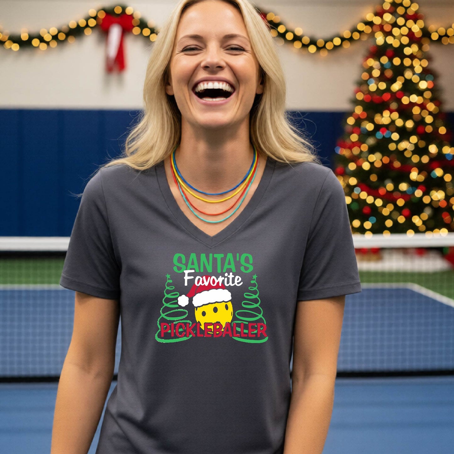 A woman smiling in a gym wearing a dark gray V-neck t-shirt with a 'Santa's Favorite Pickleballer' graphic on the front, with holiday decorations and a Christmas tree in the background.