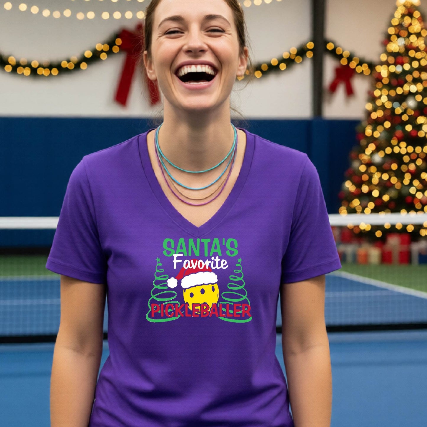 A woman smiling in a gym wearing a purple V-neck t-shirt with a 'Santa's Favorite Pickleballer' graphic on the front, with holiday decorations and a Christmas tree in the background.