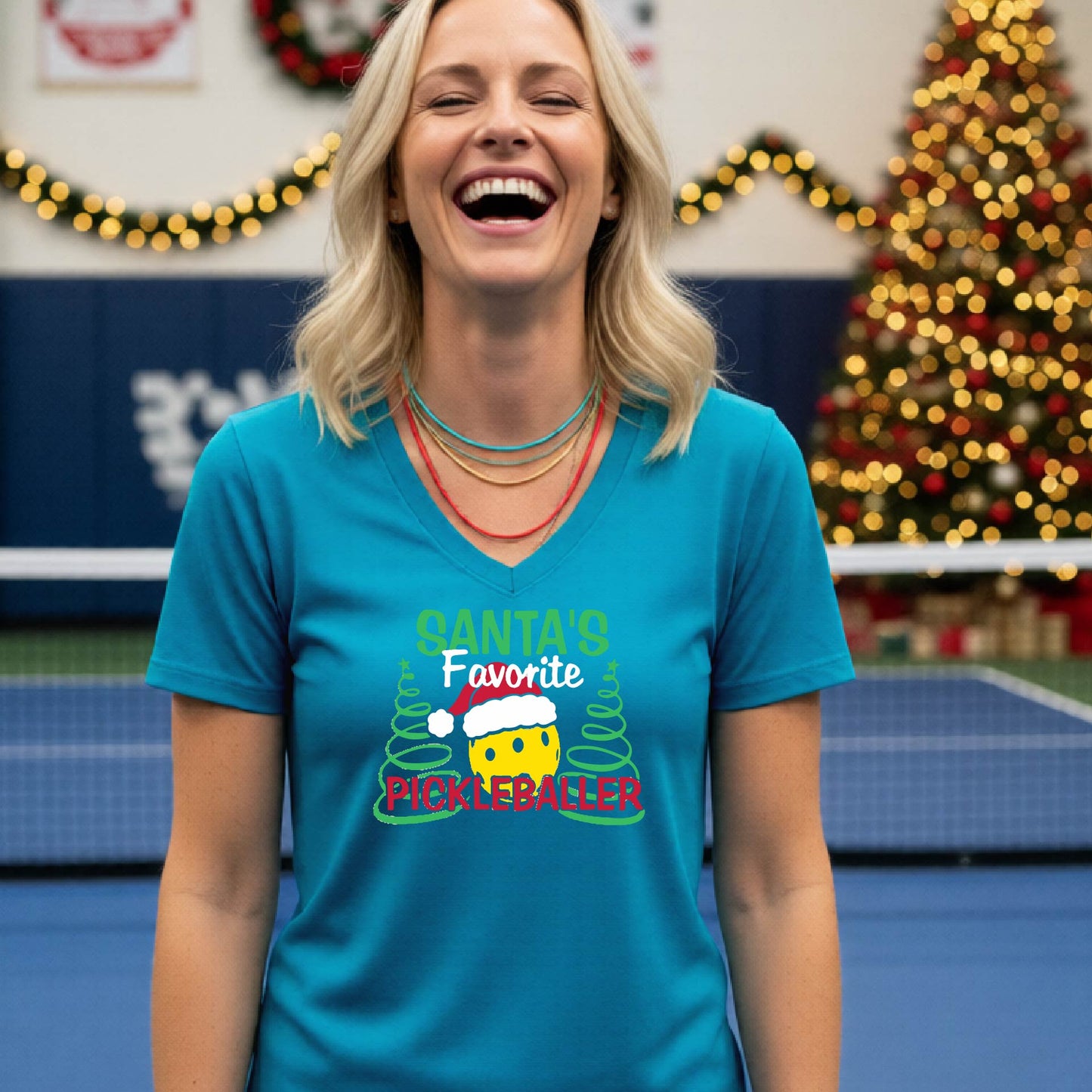 A woman smiling in a gym wearing a teal V-neck t-shirt with a 'Santa's Favorite Pickleballer' graphic on the front, with holiday decorations and a Christmas tree in the background.