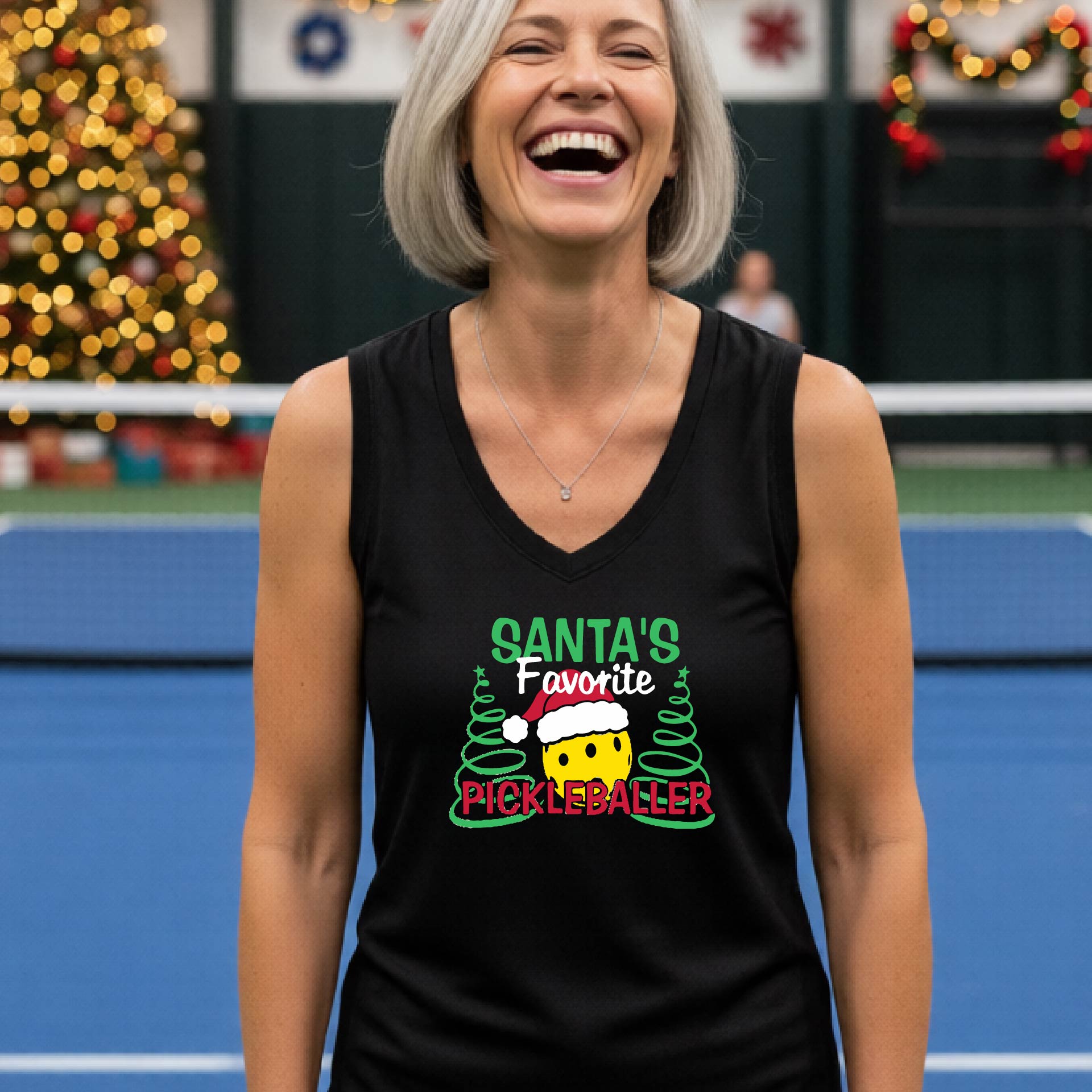 A woman laughing on a blue pickleball court wearing a black sleeveless tank top with a festive graphic that reads 'Santa's Favorite Pickleballer' with a pickleball character and Christmas trees.