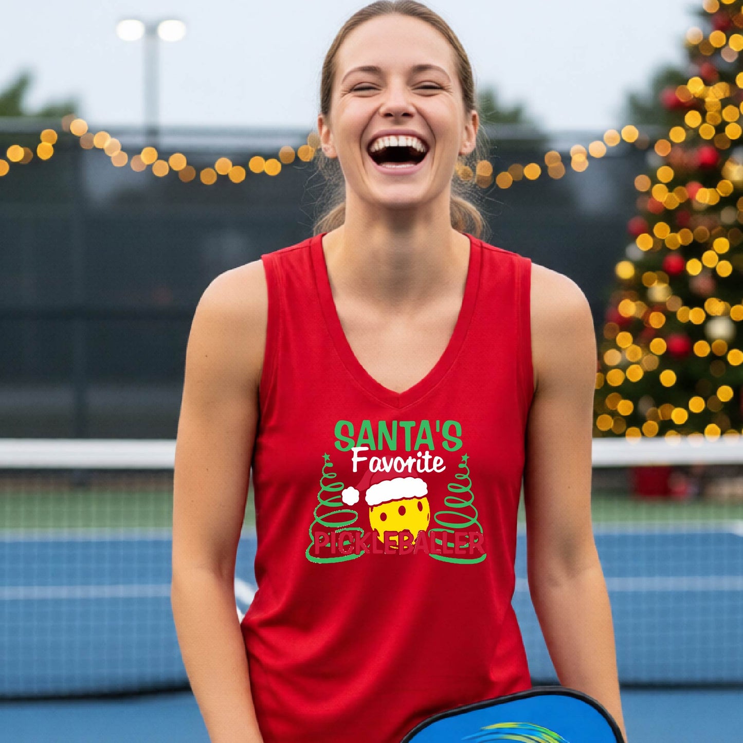A woman laughing on a blue pickleball court wearing a right red sleeveless tank top with a festive graphic that reads 'Santa's Favorite Pickleballer' with a pickleball character and Christmas trees.