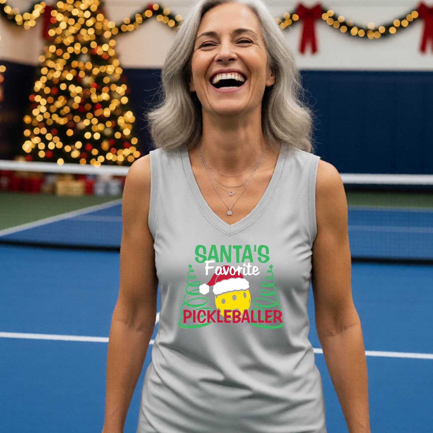 A woman laughing on a blue pickleball court wearing a silver gray sleeveless tank top with a festive graphic that reads 'Santa's Favorite Pickleballer' with a pickleball character and Christmas trees.