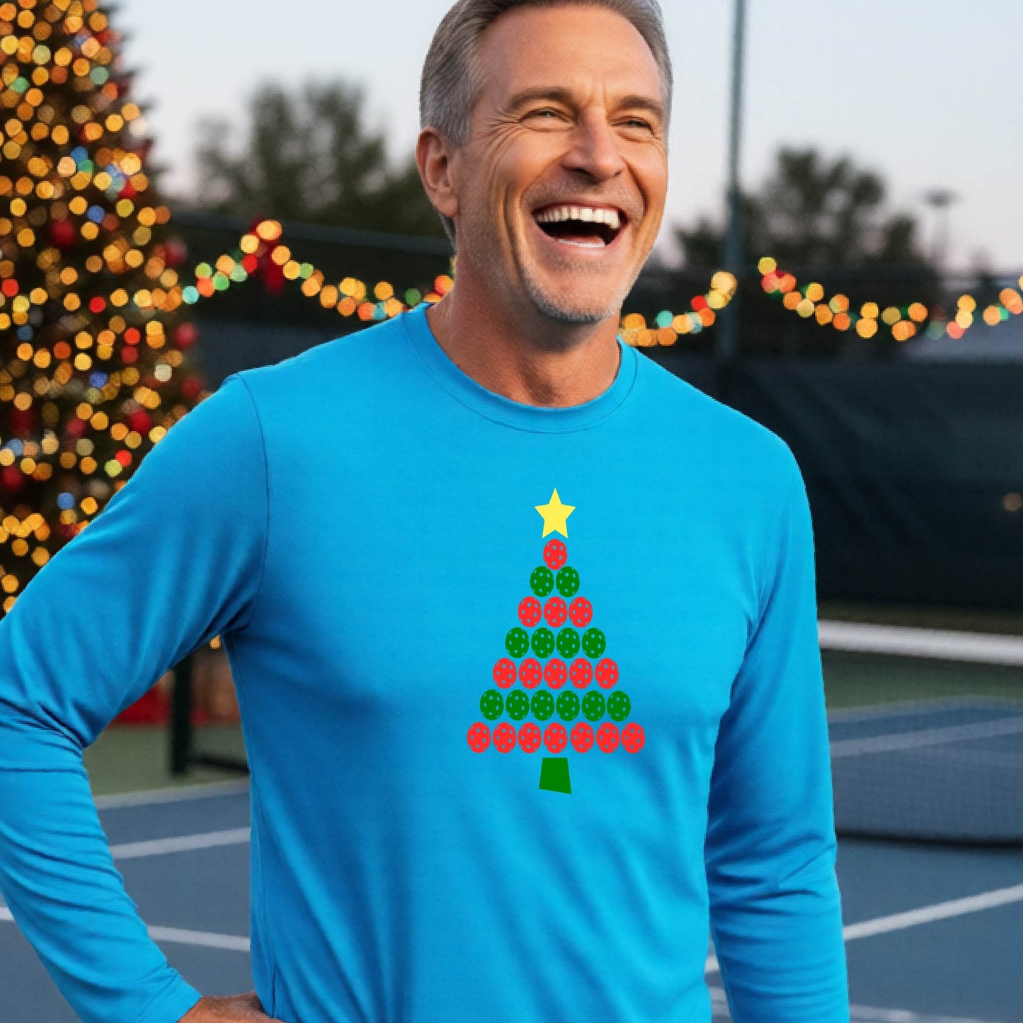 A smiling man wearing a dark bright blue long-sleeve shirt with a Christmas tree made of red and green pickleballs and a yellow star, standing on a pickleball court with holiday lights in the background.